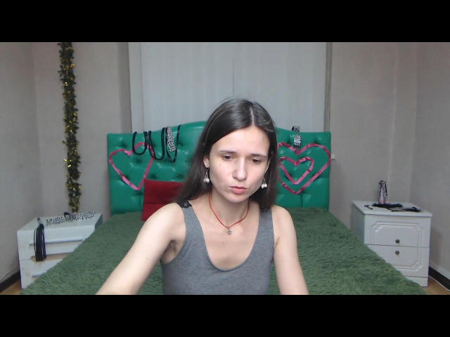 MartaLeee with long brown hair, gray tank top, and red necklace, pouting her lips, sitting on green bed with pink heart-stitched headboard, white nightstands, and Christmas decor.