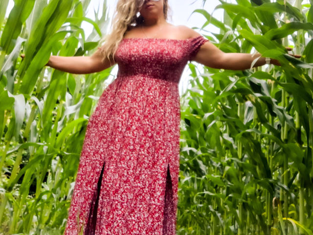 JuliaFrancaise stands in a cornfield, arms out, wearing a red floral off-shoulder dress with a slight slit, blonde hair, medium tits, and a curvy ass.