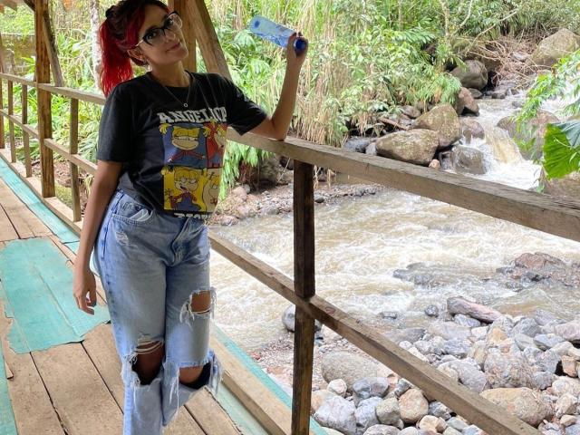 JaneRoa with red hair, glasses, black tee, and ripped jeans, standing on wooden bridge, holding phone, leaning on railing, showing her ass through torn jeans.