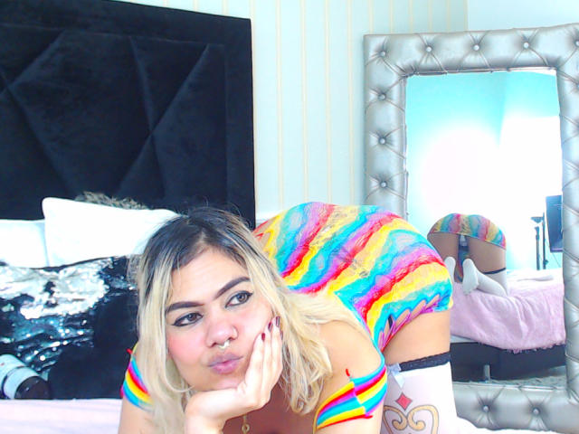 TarahDustin with blonde hair, rainbow-striped dress, and white thigh-high socks, pouting on bed, butt in mirror, black headboard, and pink bed in background.
