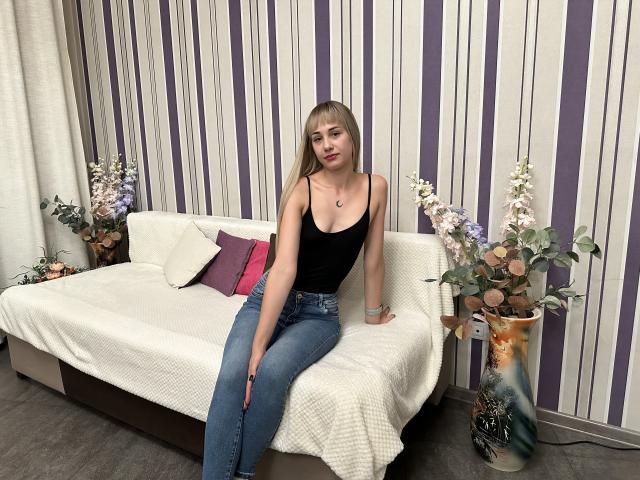 LunaBell with long blonde hair, black tank top, showing small tits, and blue jeans, sits on a white-covered couch, hands on her thighs, looking at the camera.