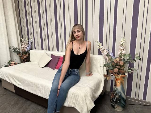 LunaBell with long blonde hair, small tits in a black tank top, and blue jeans, sitting on a white-covered sofa, right arm resting on the sofa, left hand on knee.