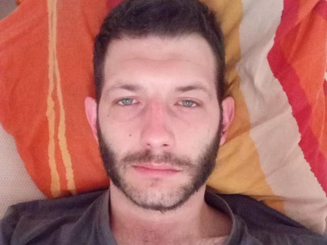 Aure69 ha una barba e dei baffi corti e scuri, occhi verdi e un labbro leggermente diviso. È sdraiato su un cuscino colorato, indossando una maglietta grigia..