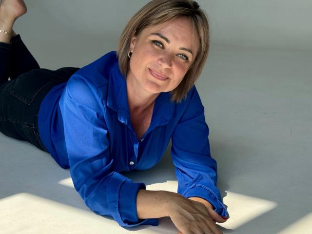 SnezhanaRells, lying on her stomach, wearing a blue shirt, black jeans, and a bracelet, with short blonde hair, smiling, hands in front, legs bent at the knees.