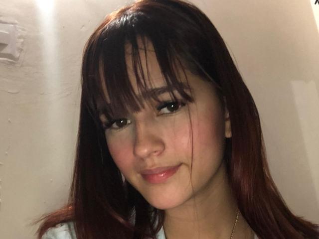 MelodyMansson has brown hair with bangs, a cute smile, and a slight blush on her cheeks. Her brown eyes are framed by long lashes. She's wearing a white top.