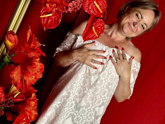 SnezhanaRells con uñas rojas, vestido blanco de encaje con hombros descubiertos, manos en el pecho, sonriendo, rodeada de lirios rojos y naranjas, anillo visible, cabello castaño corto, fondo rojo..