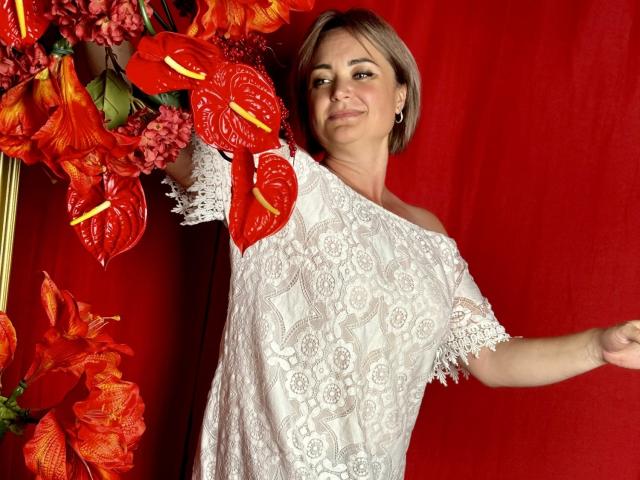 SnezhanaRells with short brown hair, white lace shirt, standing to the right, smiling, arms raised, red and orange orchids to the left, red background.