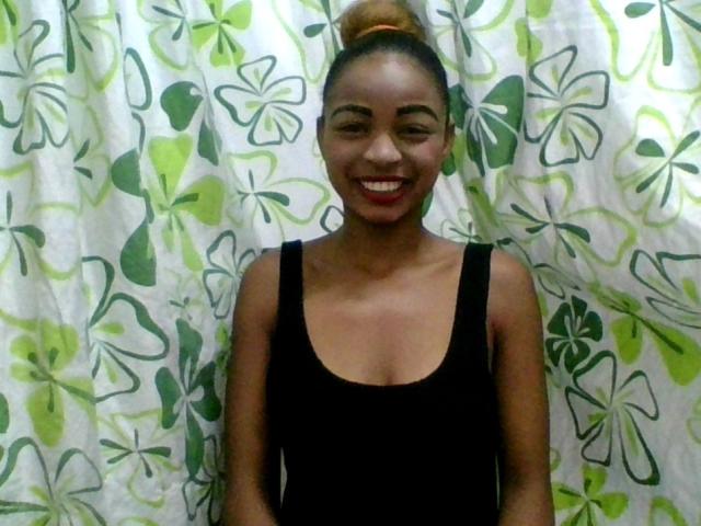 AlexaChan, smiling, with brown skin, red lipstick, black tank top, small boobs, and her hair in a bun, standing in front of a green and white floral curtain.
