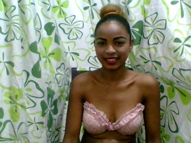 AlexaChan with brown skin, her tits in a pink, ruffled bra, smiling, red lipstick, hair in a bun, standing in front of green and white floral curtains.