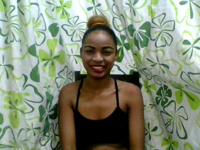 AlexaChan with dark skin, red lipstick, and a black sports bra, smiling with her brown hair in a top bun, standing in front of green and white patterned curtains.