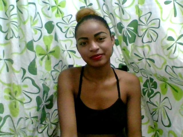 AlexaChan with tan skin, red lipstick, black sports bra, and hair in a bun, standing in front of a green and white floral curtain, looking at the camera.