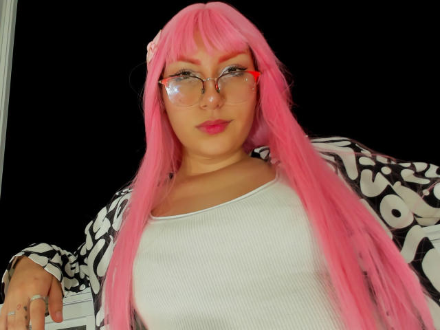LovelyLoren with pink hair, glasses, white ribbed top, black and white patterned jacket, pink lipstick, and a small flower in hair, showing a slight smile.