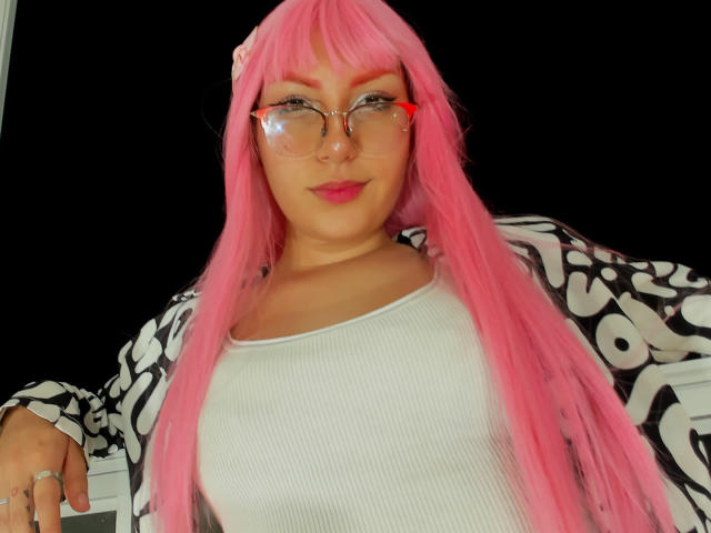 LovelyLoren has pink hair, glasses, and a white ribbed top with a black and white patterned jacket. She's smiling with pink lipstick, showing her tits slightly.