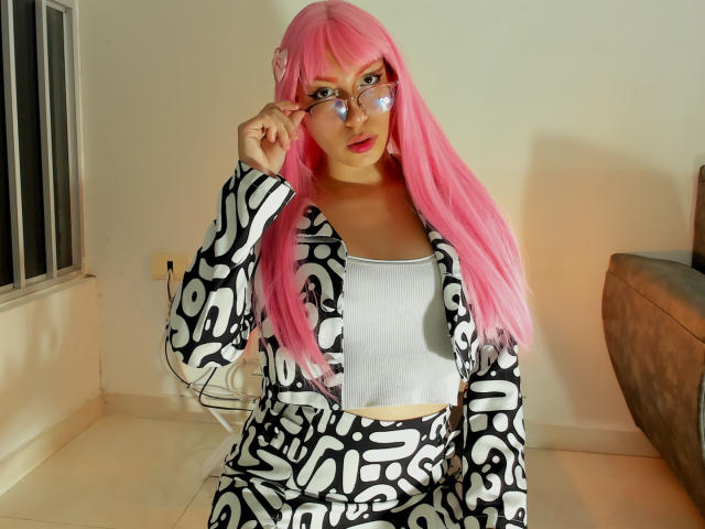 LovelyLoren with pink hair, wearing a black and white patterned outfit, white crop top, and glasses, touching her glasses, sitting on a floor, showing her tits.