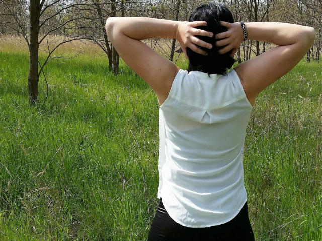 Zaubermaus83-hot with arms raised, black hair, white sleeveless top, black pants, standing in grassy field, hands on head, wearing black bracelet, back to camera.