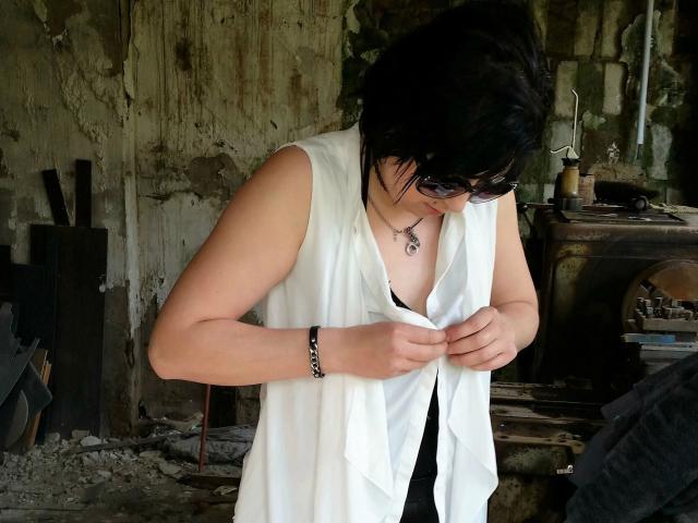 Zaubermaus83-hot with black hair, wearing sunglasses, white sleeveless vest, black top, black bracelet, and necklace, buttoning vest, showing tits, in a dirty, rusty room.