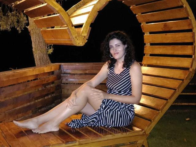 AddictedToUTS with curly dark hair, wearing a black-and-white striped dress, sits barefoot on a wooden, curved bench at night, legs crossed, smiling softly. Her hands rest on her knees.