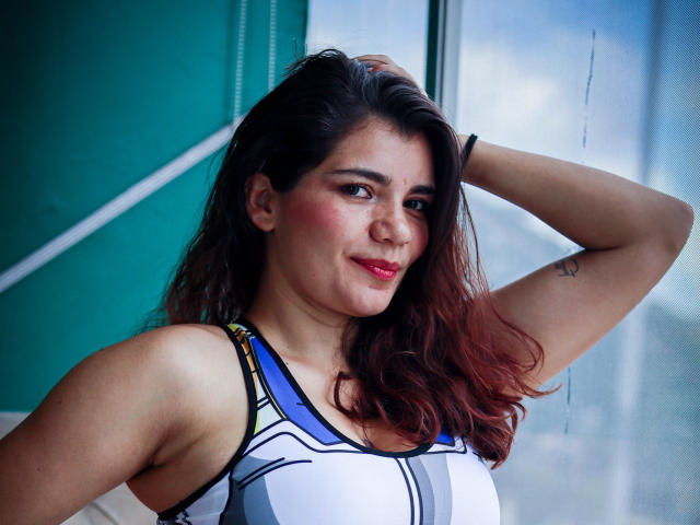 BulmaHot with dark brown hair, pink blush, and red lipstick, wearing a white and blue sports bra, arm raised, smiling, small tattoo on right arm, medium tits.