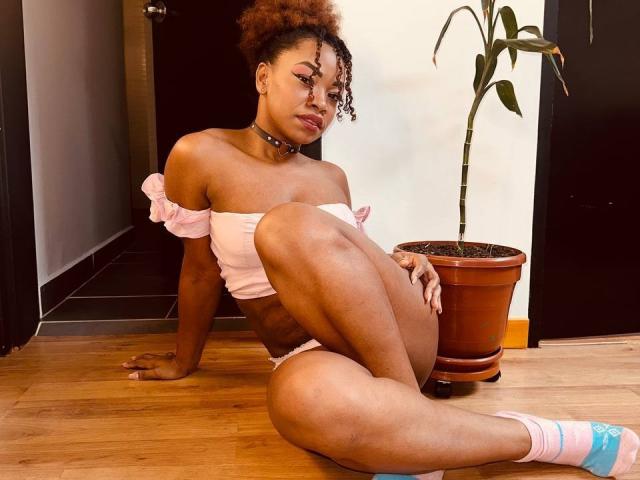 DannaBanks, con cabello castaño rizado, una blusa rosa con un hombro descubierto y un collar, sentada en el suelo de madera, con las piernas separadas, calcetines rosas, una pequeña maceta marrón con una planta, y una puerta negra en el fondo..