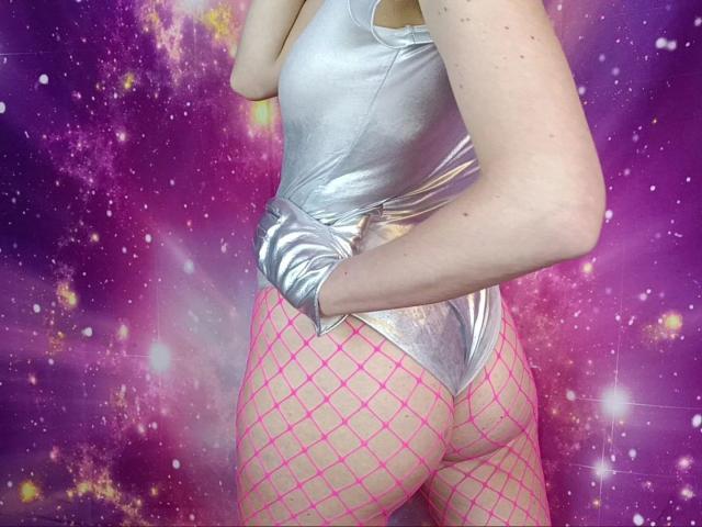 Lynn-Tonic-hot wearing a shiny silver top, pink fishnet stockings, and her ass is exposed. Her hand is in her top, showing her butt. She's standing with her back.