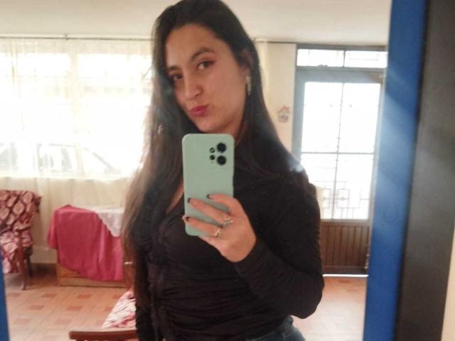 VenusSweetPalmer with long dark hair, wearing a black long-sleeve top, taking a mirror selfie, showing her tits, holding a mint-green phone, in a brightly lit room.