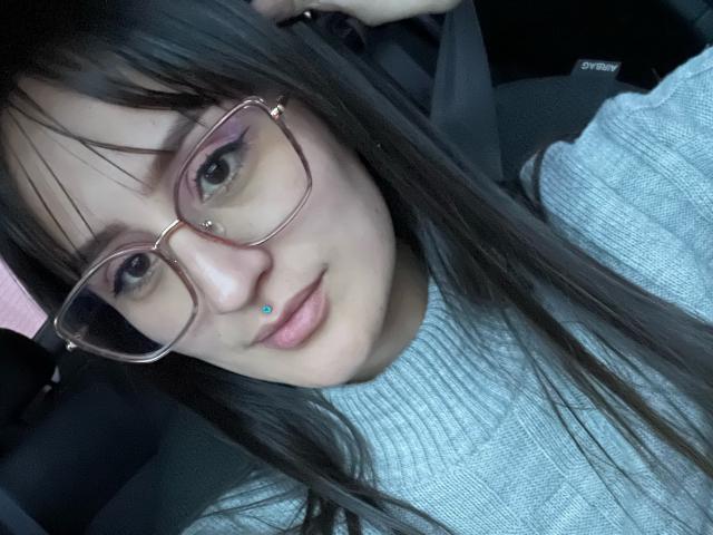 NinaBryons with long black hair, glasses, nose piercing, light gray sweater, and slight smile, looking at camera in car. Her tits are not visible.