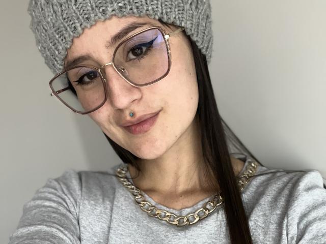 NinaBryons with glasses, gray beanie, nose piercing, and chain necklace, wearing a gray top, smiling slightly, long brown hair, light skin, and subtle makeup.