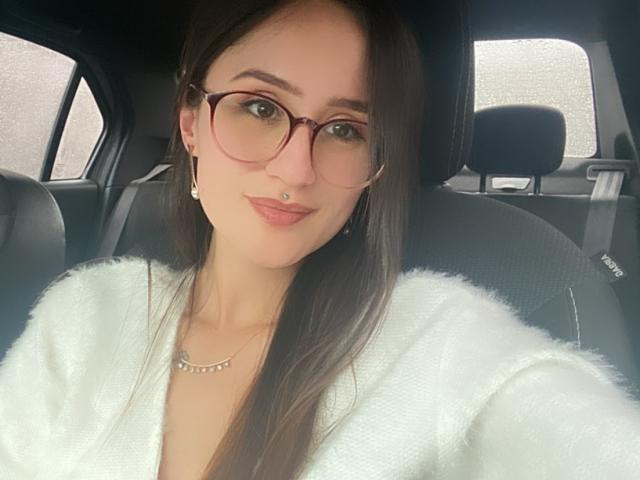 NinaBryons with glasses, white fuzzy sweater, nose ring, and long brown hair, sitting in a car. She's smiling slightly, wearing a necklace, with a calm expression.