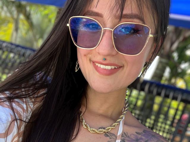 NinaBryons with blue reflective sunglasses, a nose piercing, a chain necklace, and tattoos, smiling with dark hair, white top, and hoop earrings, showing her tits in the photo.