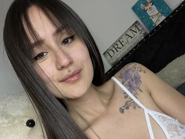 NinaBryons has long brown hair, brown eyes, and a blue nose piercing. She's wearing a white lace top, showing a purple tattoo on her right shoulder, and smiling.