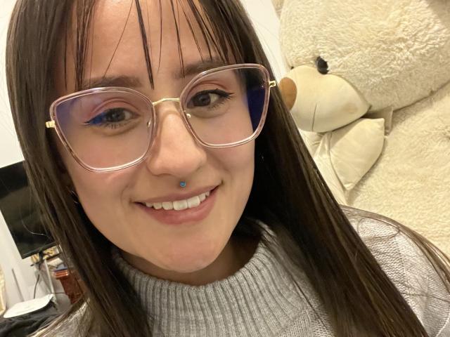 NinaBryons with long brown hair, glasses, nose piercing, blue eyeliner, smiling, gray turtleneck, white fluffy teddy bear in background, standing in well-lit room.