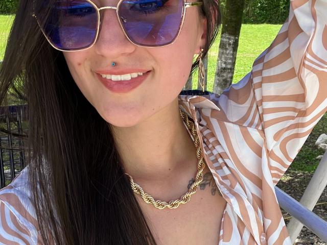 NinaBryons with blue-tinted aviator sunglasses, brown-striped shirt, gold chain necklace, nose piercing, smiling, long brown hair, green grass, and trees in the background. Boobs not visible.