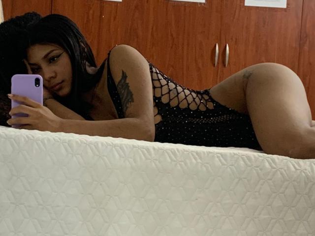 NaomiPortia lying on a bed, wearing a black fishnet bodysuit, showing her ass, holding a purple phone, with a tattoo on her arm, in a bedroom.