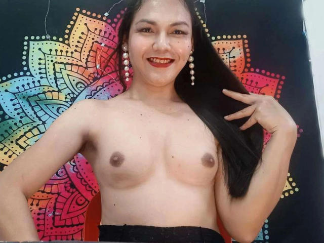 AsianCutieModel-hot with long black hair, smiling, wearing pearl earrings, topless, showing small tits, standing against a colorful, mandala-patterned background, hand touching her hair.