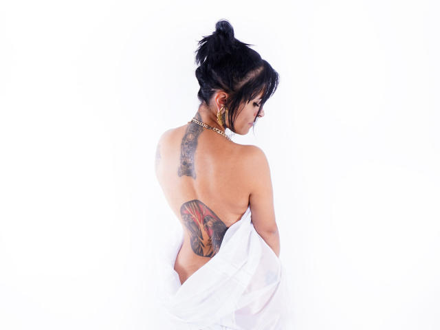 AmelieBlake with black hair in a bun, tattooed back, naked except for a white shirt, gold necklace, and earrings, facing away, showing her tits and ass.