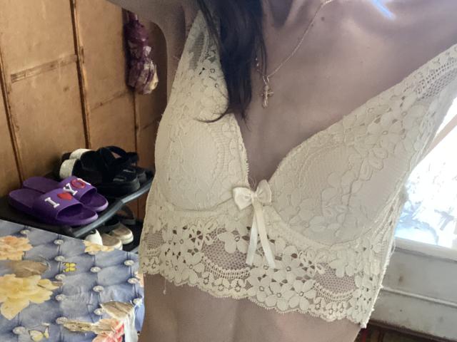 Kabblasha's small tits in a white lace bralette with a bow, her neck adorned with a cross necklace, in a dimly lit room with floral bedcovers.