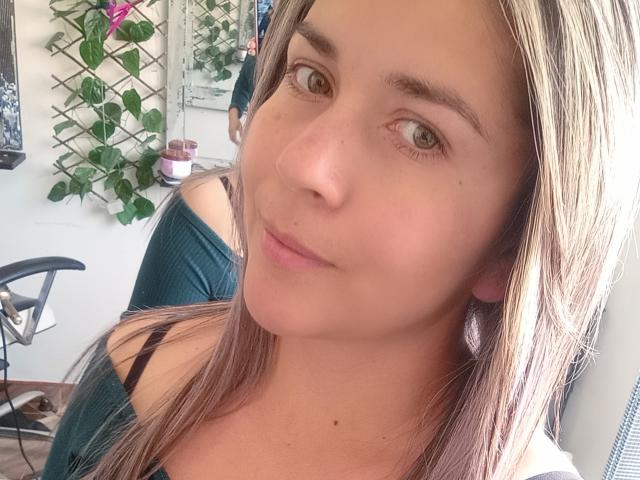 DianaAme with light brown hair, green eyes, and a slight smile, wearing a green off-shoulder top, showing her black bra strap and small pink earrings.