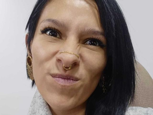 AmelieBlake has black hair, a gold nose chain, a ringed nose, and is pouting her lips with a slight frown, wearing a gray fur collar and ornate earrings.