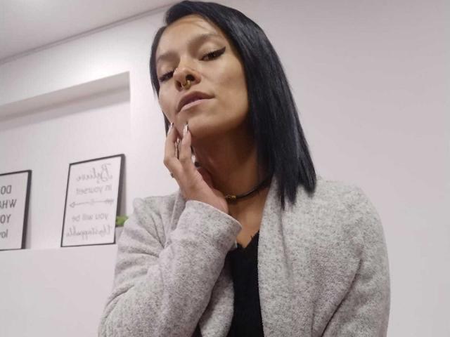 AmelieBlake has black hair, a nose ring, and a choker. She's wearing a gray cardigan, touching her cheek, with a confident, slightly pouty expression.
