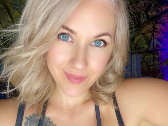 Misstyk has blonde hair, blue eyes, and a cute smile. She's wearing a black top with visible tits and a tattoo on her chest. Her makeup is subtle and natural.