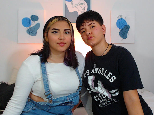 CamilleAndSophie with dark hair, pouty lips, and white top in blue overalls, and Sophie with short black hair, nose ring, and black tee, standing in a room.