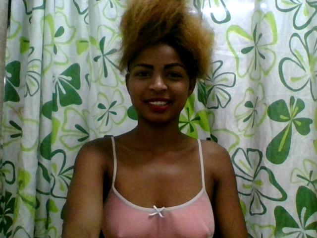 AlexaChan with medium brown skin, curly afro, red lipstick, smiling, wearing pink camisole with white straps, small white bow, standing in front of green and white floral curtain.