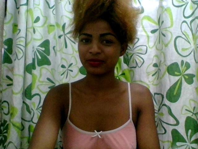 AlexaChan with dark skin and curly blonde hair, wearing a pink camisole, has small tits, red lipstick, and a cute smile, standing in front of green floral curtains.