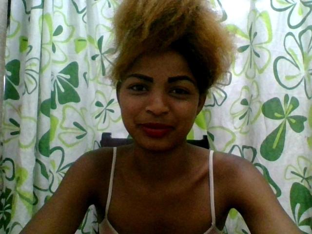 AlexaChan with light brown, puffy Afro, red lipstick, and white spaghetti strap top, smiling slightly, showing her small, perky tits, sitting in front of green floral curtains.