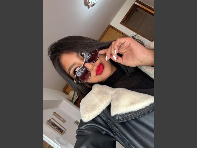 CarlaRusso with dark hair, red lipstick, and black leather jacket with white fur collar, wearing aviator sunglasses, adjusting them with her right hand.