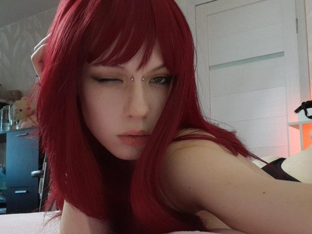 MonyBabe with red hair, winking, has a nose piercing and lip piercing, looking seductively at the camera, showing her bare shoulder. She's a pale-skinned, cute, sexy chick.