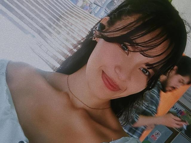 SusanKartier with wet black hair, red lipstick, off-shoulder white top, small bare boobs, smiling, black earbuds, thin necklace, blurred man in background, daytime, casual selfie.