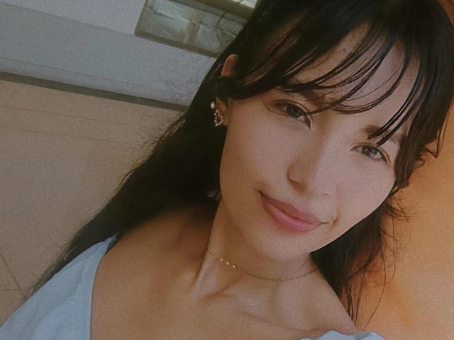 SusanKartier has a cute smile, light skin, black bangs, and wears a light blue top, showing her small tits and a thin gold necklace. She's looking at the camera.
