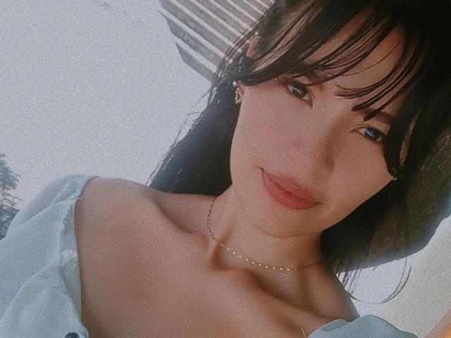 SusanKartier with dark brown hair, bangs, and a subtle smile, wearing a light blue off-shoulder top, showing her bare shoulders and a delicate necklace.