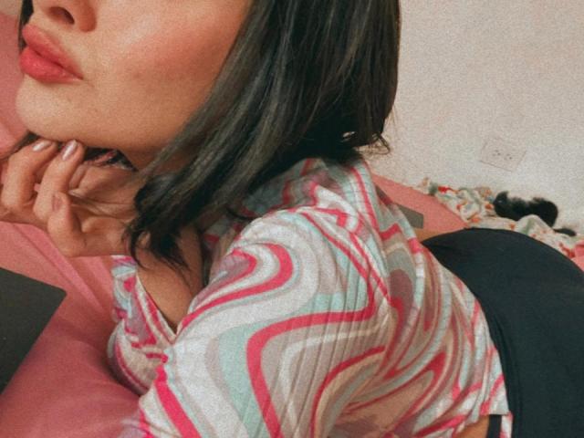 IsabellaPalacio with pouty lips, lying on pink bed, wearing a striped shirt, black skirt, and white nails, showing her ass and hands near her face.